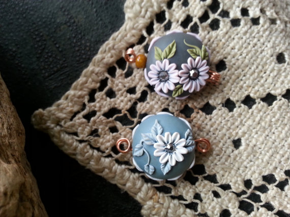 2 handcrafted focal beads, the first is steel blue background with flowers & stitching in bright white, and foliage in taupe and the second is pale pink with green foliage on a dark grey background.